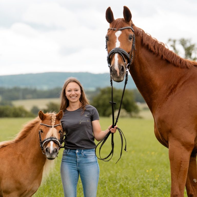 Frau steht mit einem kleinen und einem großen Pferd auf einer Wiese.