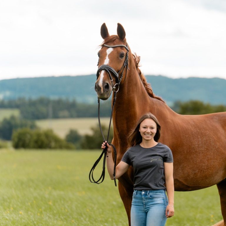 Frau steht neben einem braunen Pferd auf einer Wiese mit grünen Hügeln im Hintergrund.